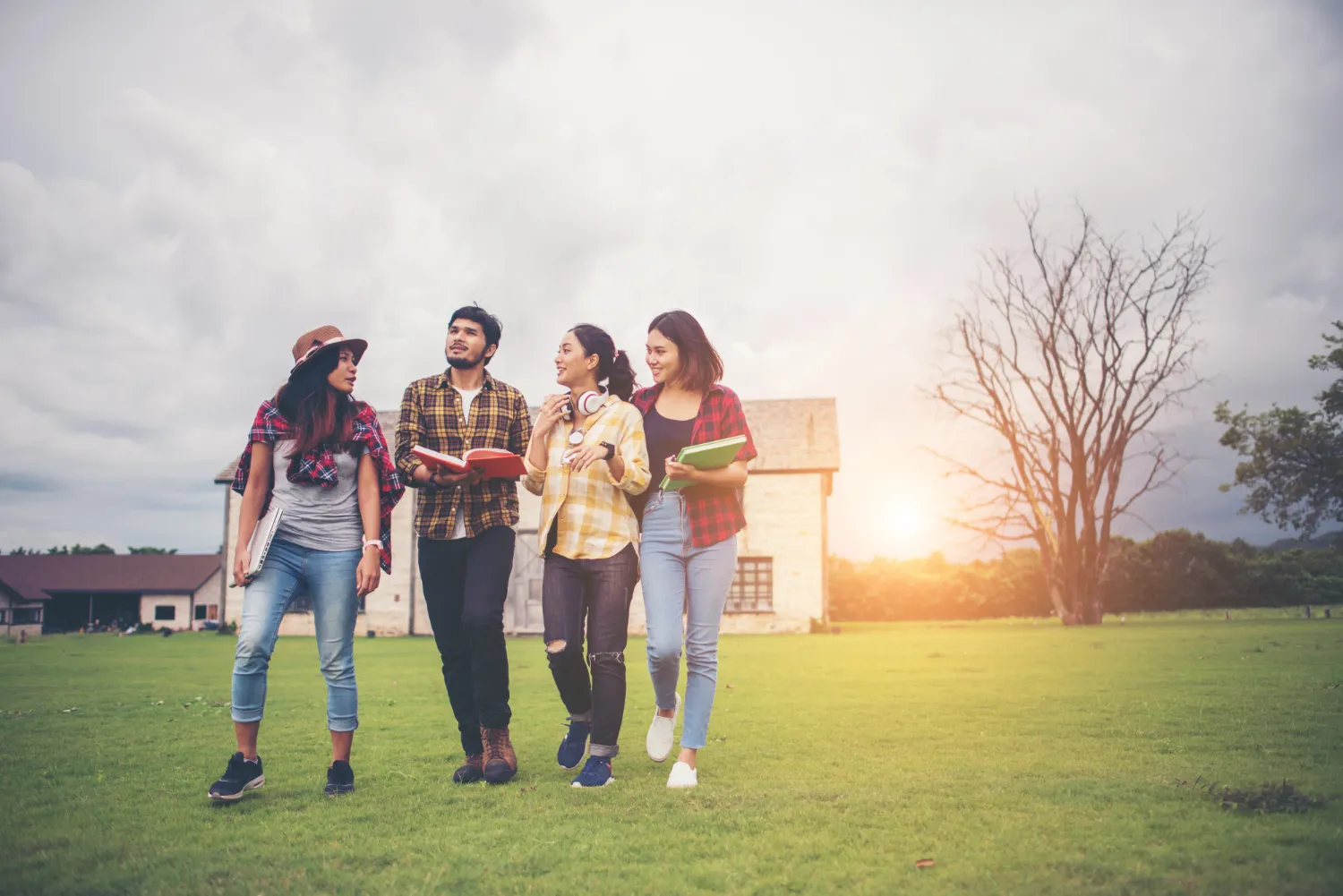 Four young adults walking on grass outdoors carrying books and chatting near a building at sunset.
